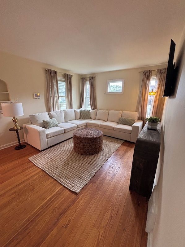 Ein Wohnzimmer mit einem Ecksofa, einem runden Hocker, einer Stehlampe und einem flachbildfernseher, der an der Wand montiert ist.