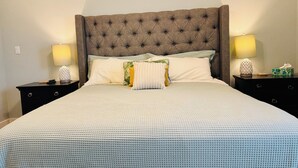 Una cama bien hecha con cabecera capitonada, dos mesitas de noche con lámparas, y una mesita de noche con un dispensador de pañuelos.