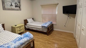 Quarto com duas camas, ventoinha no teto, televisão fixada na parede e janela com persianas.