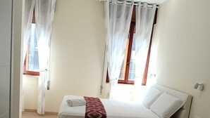 Ein Schlafzimmer mit einem Bett, einem Schreibtisch und einem Stuhl. Vor den Fenstern hängen Vorhänge.