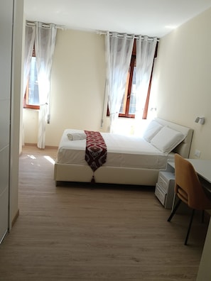 Ein Schlafzimmer mit einem Bett, einem Schreibtisch und einem Stuhl. Vor den Fenstern hängen Vorhänge.