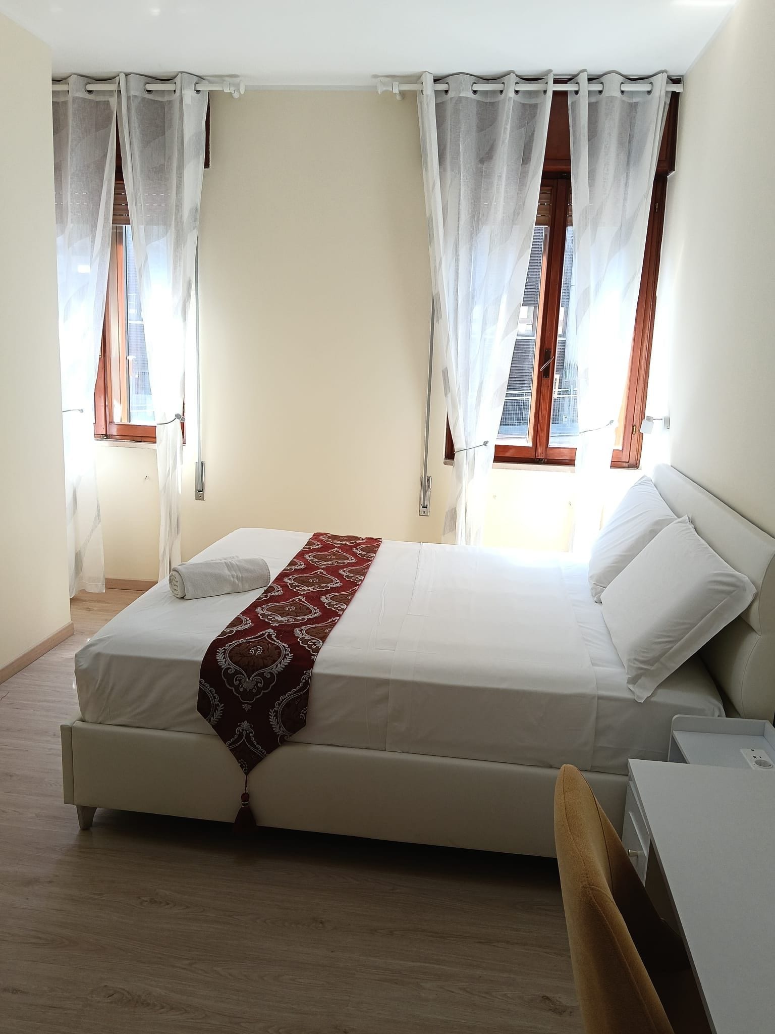Une chambre à coucher avec un lit, deux fenêtres avec des rideaux, une table de chevet et une chaise.