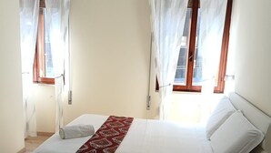 Ein Schlafzimmer mit einem Bett, zwei Fenstern mit Vorhängen, einem Nachttisch und einem Sessel.