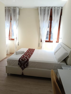 Ein Schlafzimmer mit einem Bett, zwei Fenstern mit Vorhängen, einem Nachttisch und einem Sessel.