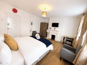Quarto de hotel com cama, poltrona, lareira, televisão e relógio na parede.