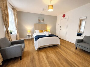 Ein Hotelzimmer mit einem Bett, zwei Sesseln, einem Nachttisch mit Lampe, einer Wanduhr und einem Spiegel.