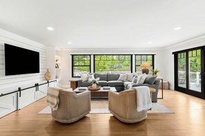 A modern living room with a large flat-screen TV, a grey sofa set, wicker chairs, and a wooden coffee table. - The Creekhouse | Water Views | Lux Extended Stays (Portsmouth)