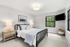 Ein Schlafzimmer mit Bett, Nachttisch, Lampe, gerahmtem Bild, Fenster und einem Fernseher.