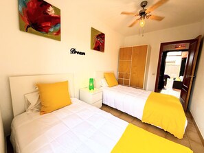 A bedroom with two beds, each with a white headboard and yellow bedding. There is a ceiling fan, a green bottle on the nightstand, and a door leading to a balcony. - Nur house villa & private pool (La Oliva)