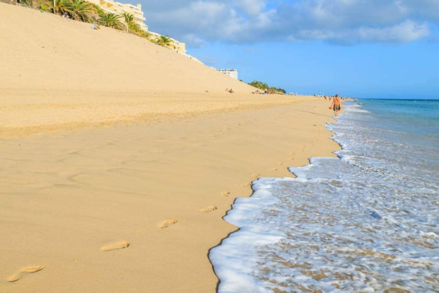 Une plage de sable avec des empreintes menant à l’eau, un ciel dégagé et un bâtiment en arrière-plan.