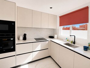 A modern kitchen with white cabinets, a black microwave, a sink, and a window with a red blind. - Alma Nomada Luxury Village & Private Pool & BBQ & ChillOut (Corralejo)