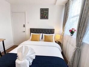 A hotel room with a bed, a bedside table, a nightstand, a lamp, a framed botanical illustration, and a window with curtains. - Double Room in East London near Aldgate East (London)