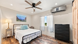 A bedroom with a bed, a nightstand, a lamp, a dresser, a ceiling fan, and a window with curtains. - Buttons on the Pedernales Casita - Riverview (Stonewall)