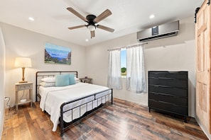 Quarto com cama, mesa de cabeceira, luminária, cômoda, ventilador de teto e janela com cortinas.