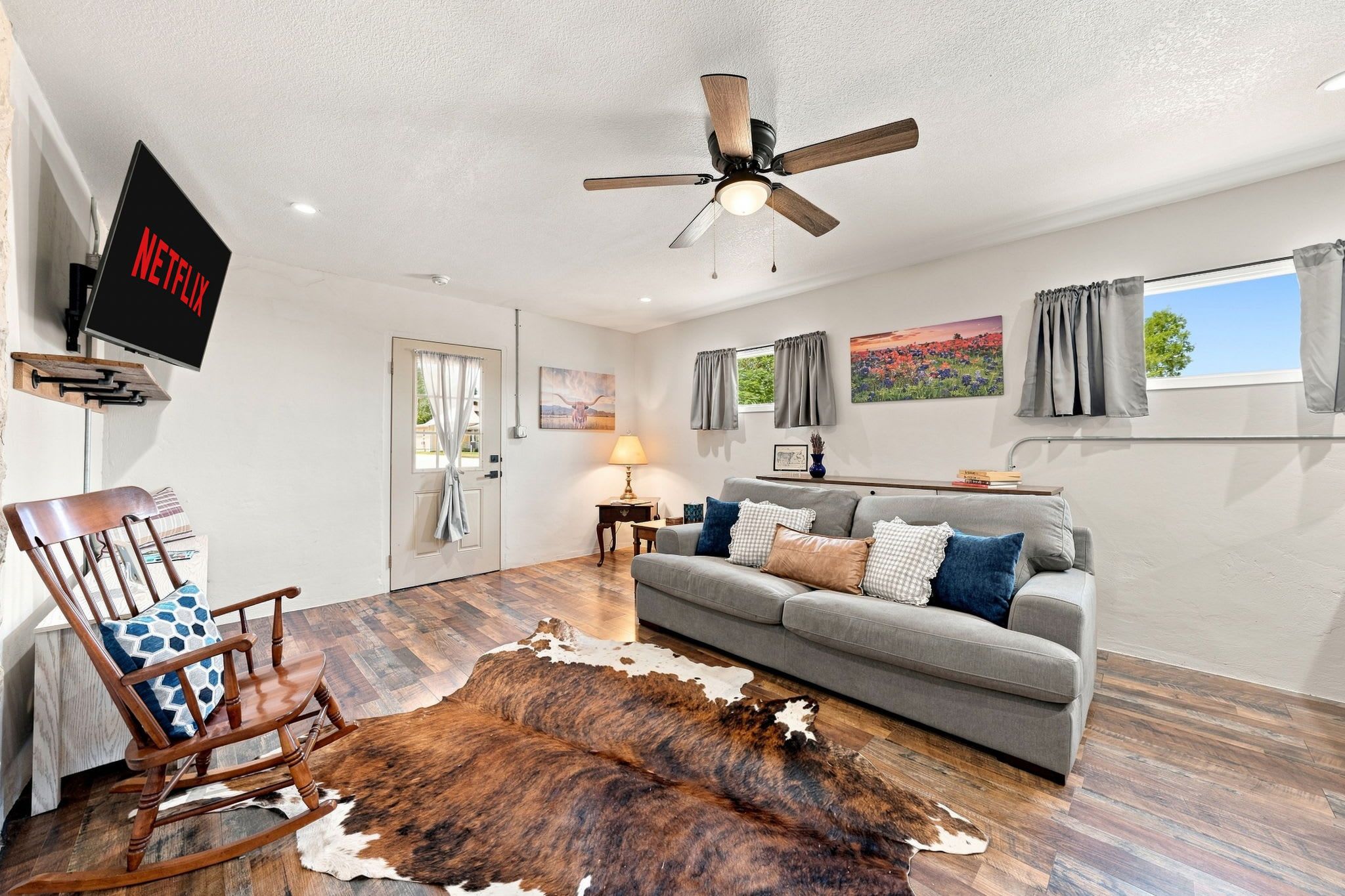A living room with a grey sofa, a wooden rocking chair, a ceiling fan, and a mounted television displaying 'NETFLIX'.