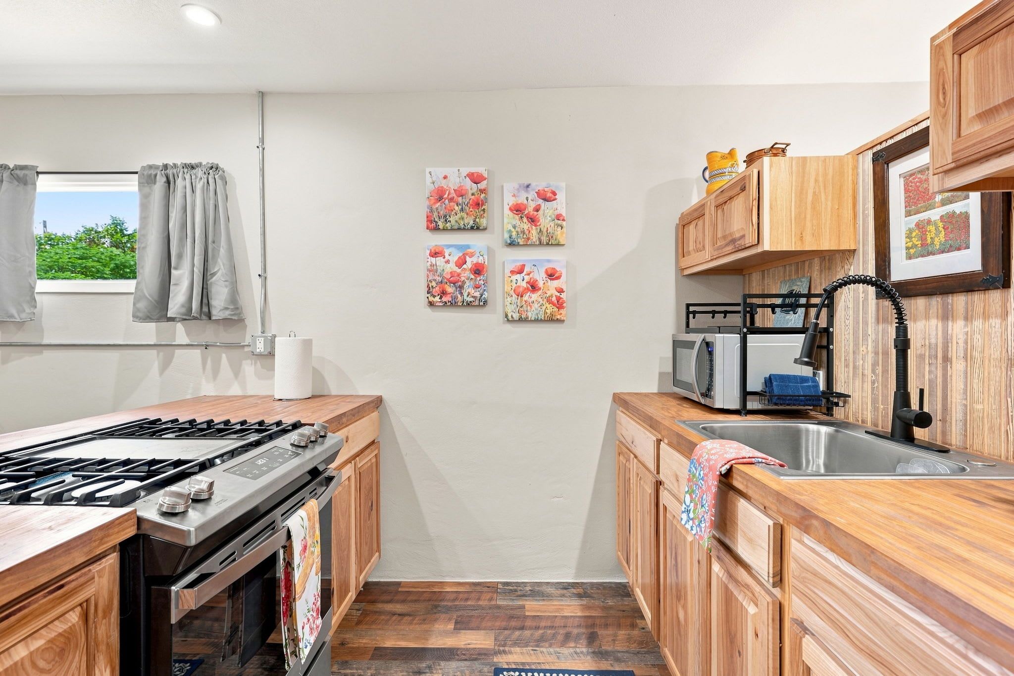 A compact kitchen with a gas stove, wooden cabinets, a microwave, and a sink. There are four floral wall art pieces and a window with curtains.
