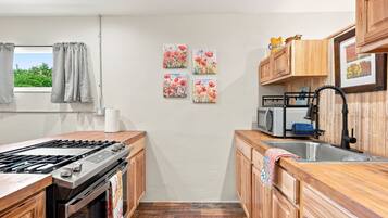 A compact kitchen with a gas stove, wooden cabinets, a microwave, and a sink. There are four floral wall art pieces and a window with curtains.