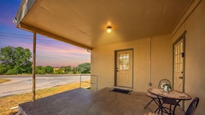 A covered porch with a concrete floor, a small table with chairs, and a door with a window. - Buttons on the Pedernales Casita - Riverview (Stonewall)