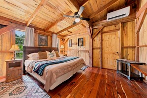 A wooden cabin interior with a bed, a ceiling fan, an air conditioning unit, and a rustic wooden door. - Saw Mill (Lodge #4) Hot Tub & 10 Mins to Main St (Fredericksburg)