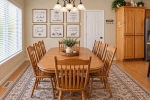 A dining area with a wooden table and chairs, a chandelier, framed welcome signs, and a kitchen area with wooden cabinets.