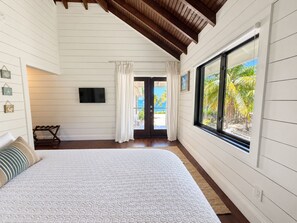 A bedroom with a large bed, a wooden ceiling, a window with a view of palm trees, and a door leading outside. - Camp Bay Beach House – Private Pool and Beachfront (Camp Bay)