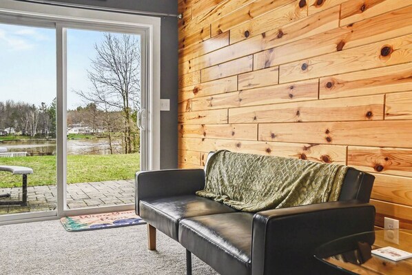 A modern living room with a black leather sofa, a wooden wall, and a glass door leading to an outdoor area.