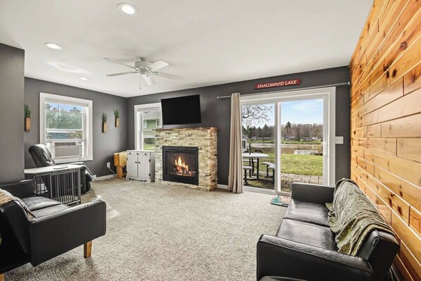 A living room with a fireplace, a ceiling fan, and a view of a lake. - Lakefront Retreat - Gladwin (Gladwin)
