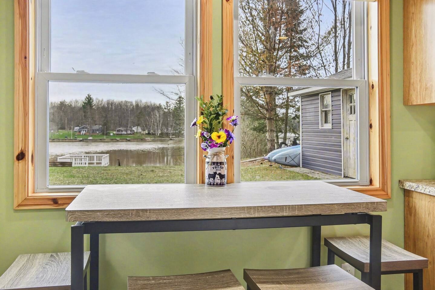 Um quarto com uma mesa e cadeiras, uma janela com vista para um lago e uma casa, e um vaso com flores na mesa.