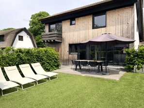 A modern building with a wooden exterior, a covered outdoor seating area with a table and chairs, and a lawn with lounge chairs. - Group Stay Near Schoorl Dunes (Schoorl)