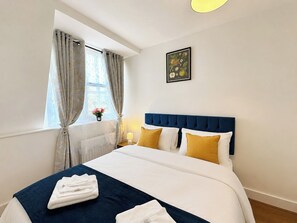 Habitación de hotel con cama, mesita de noche, ventana con cortinas y un cuadro colgado en la pared.