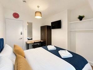 Habitación de hotel con cama, escritorio, silla, armario, televisor y reloj.