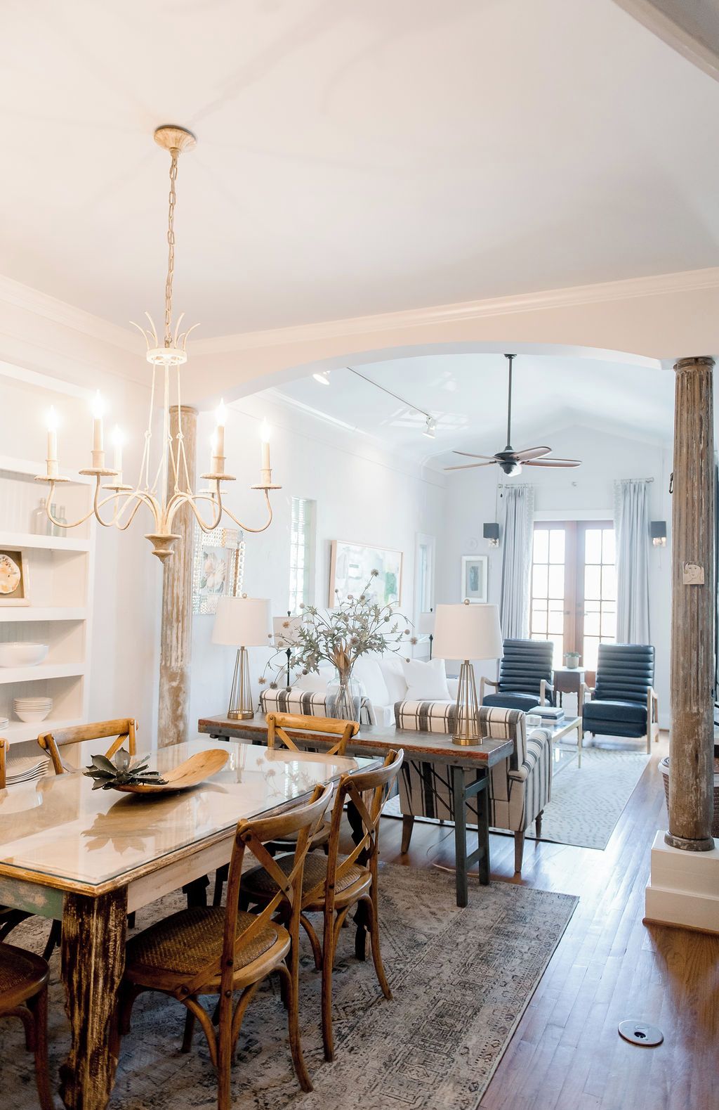 A spacious dining area with a wooden table and chairs, a chandelier, and a view into a living room with a sofa and a ceiling fan.