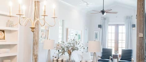 A spacious dining area with a wooden table and chairs, a chandelier, and a view into a living room with a sofa and a ceiling fan.