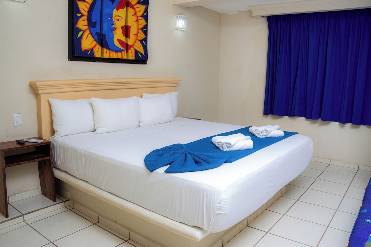 Una habitación de hotel con una cama grande, ropa de cama blanca, una colcha azul y una cortina azul.