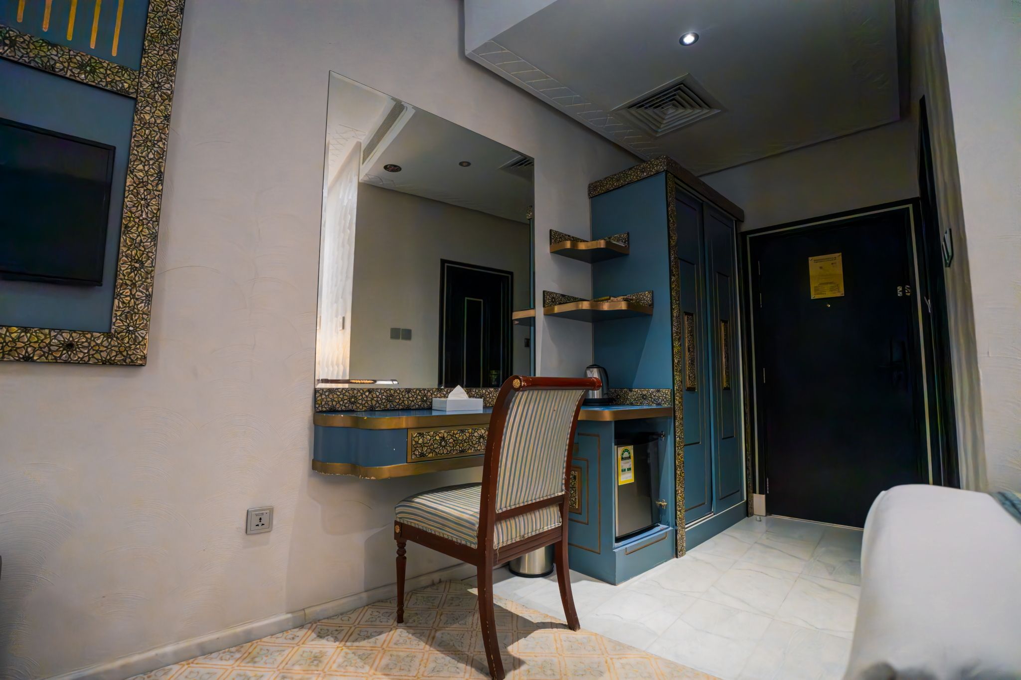 Habitación de hotel con un espejo grande, una silla de madera, una mesita con una caja de pañuelos, un armario azul y una puerta con un letrero.