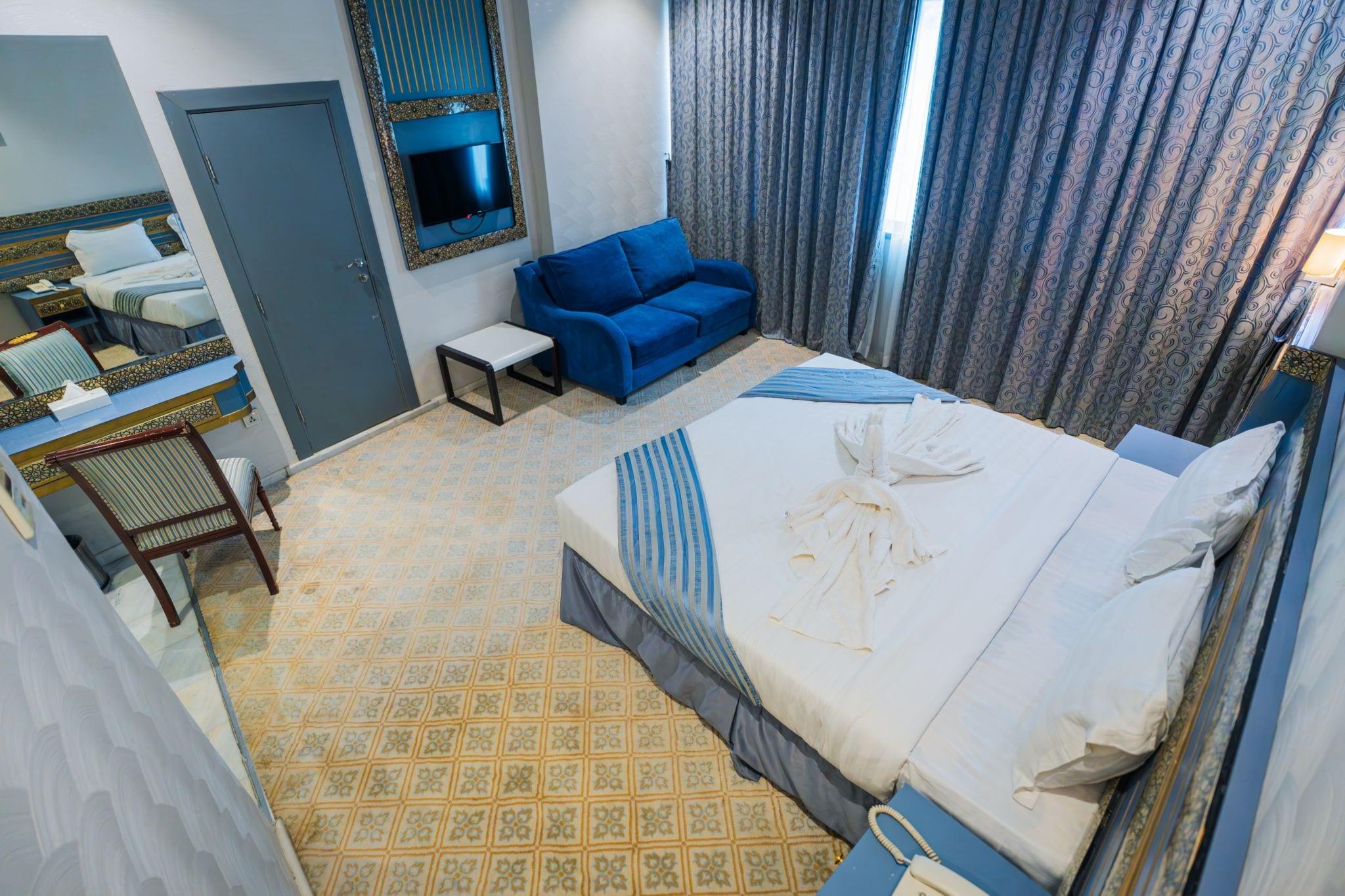 Quarto de hotel com uma cama, sofá azul, mesinha, televisão fixada na parede e piso estampado.