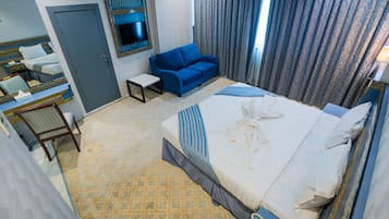 Quarto de hotel com uma cama, sofá azul, mesinha, televisão fixada na parede e piso estampado.