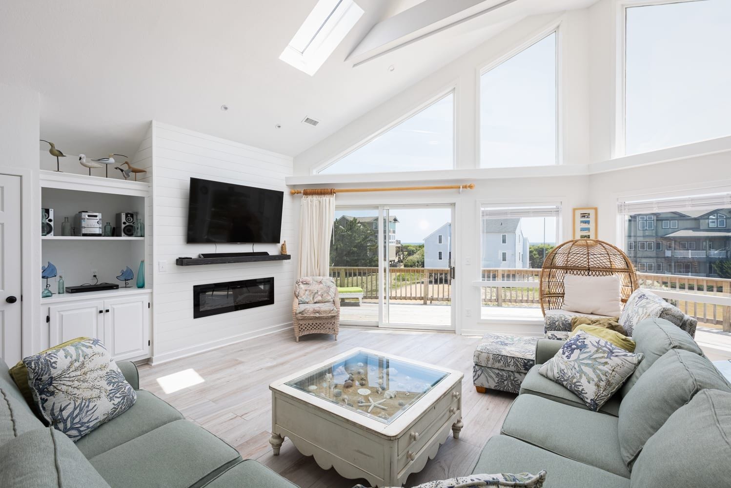 A spacious living room with a flat-screen TV, a glass coffee table, and a balcony with a wicker chair.
