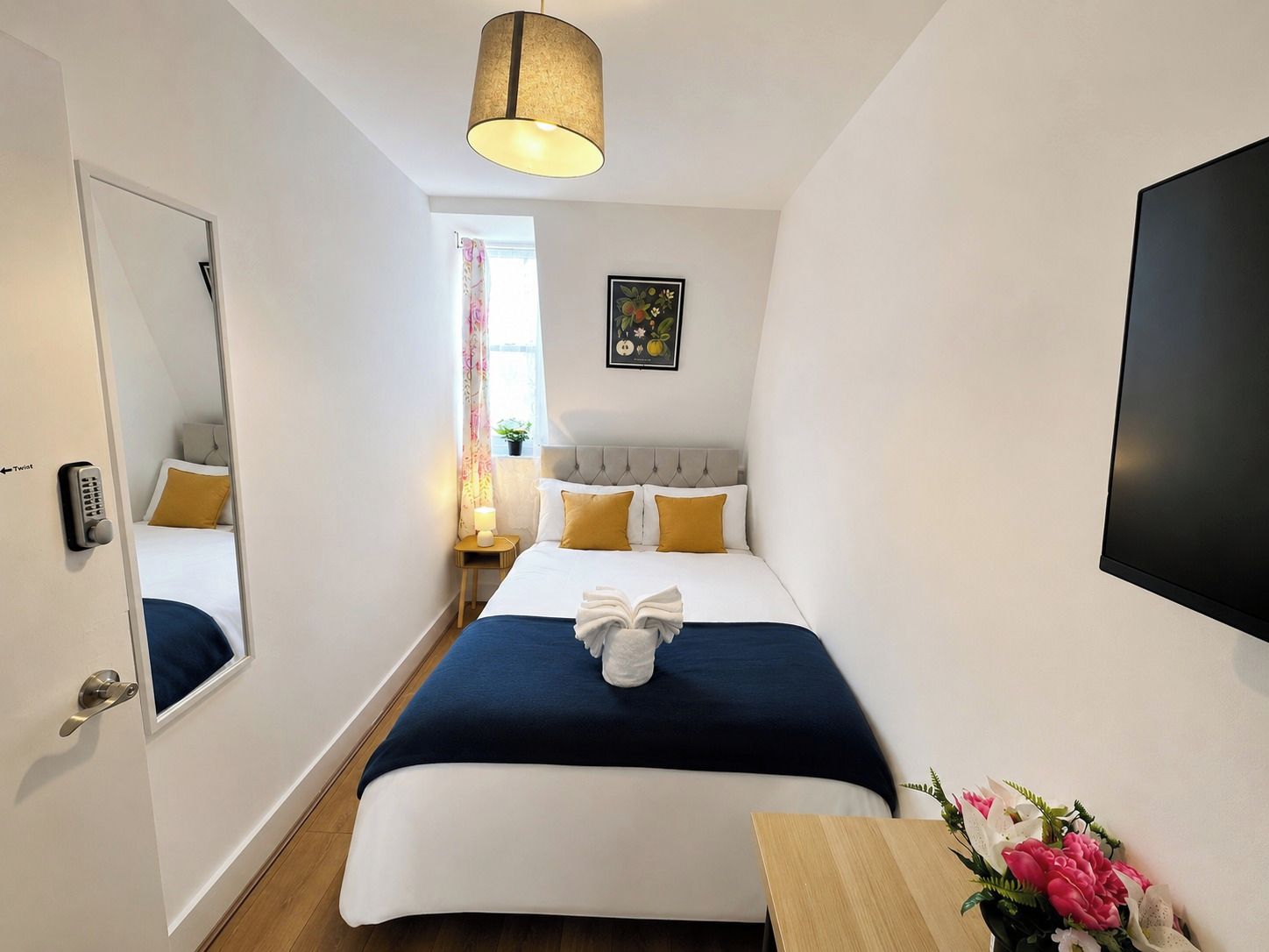Una habitación de hotel con una cama, una mesita de noche, una lámpara, un espejo y un televisor instalado en la pared.