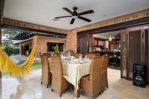An outdoor patio with a dining table set for four, surrounded by wicker chairs, a ceiling fan, and a hammock.