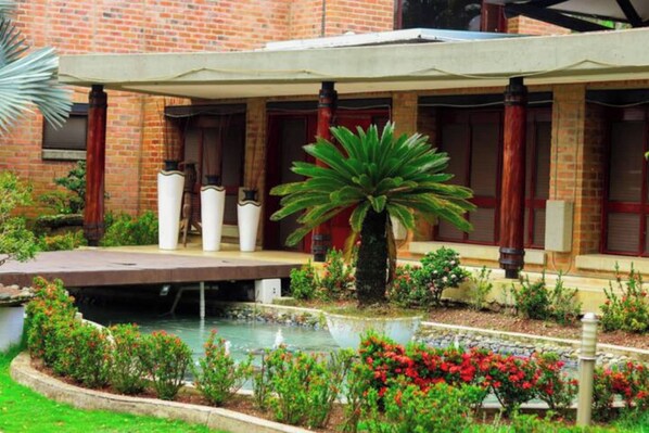 A brick building with a covered patio, a small pond, and various plants.