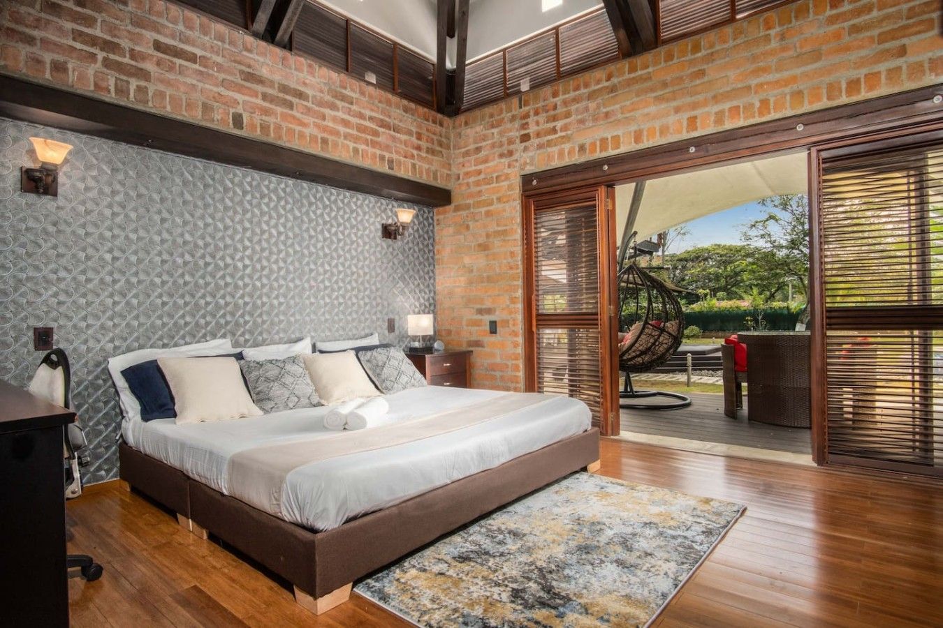 A bedroom with a large bed, a wooden floor, a patterned wall, and a view through a glass door.