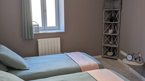 Et soveværelse med to senge, et vindue med gardiner, en radiator, en hylde med pynteting og et lille bord med en cykel og et ur.