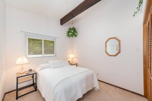 Ein Schlafzimmer mit einem Bett, zwei Nachttischen, einem Fenster mit Jalousien, einer hängenden Pflanze und einem gerahmten Bild an der Wand.