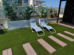 Eine Dachterrasse mit Kunstgrasanlage, zwei Liegestühlen und Blick auf die Stadtlandschaft.