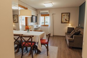 Un comedor con una mesa, sillas y un florero. Se ve un sofá y un televisor al fondo.