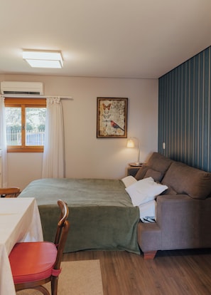 A room with a bed, a sofa, a table, a chair, an air conditioning unit, a window with curtains, a wall-mounted lamp, and a framed picture. - Belo Destino (Gramado)