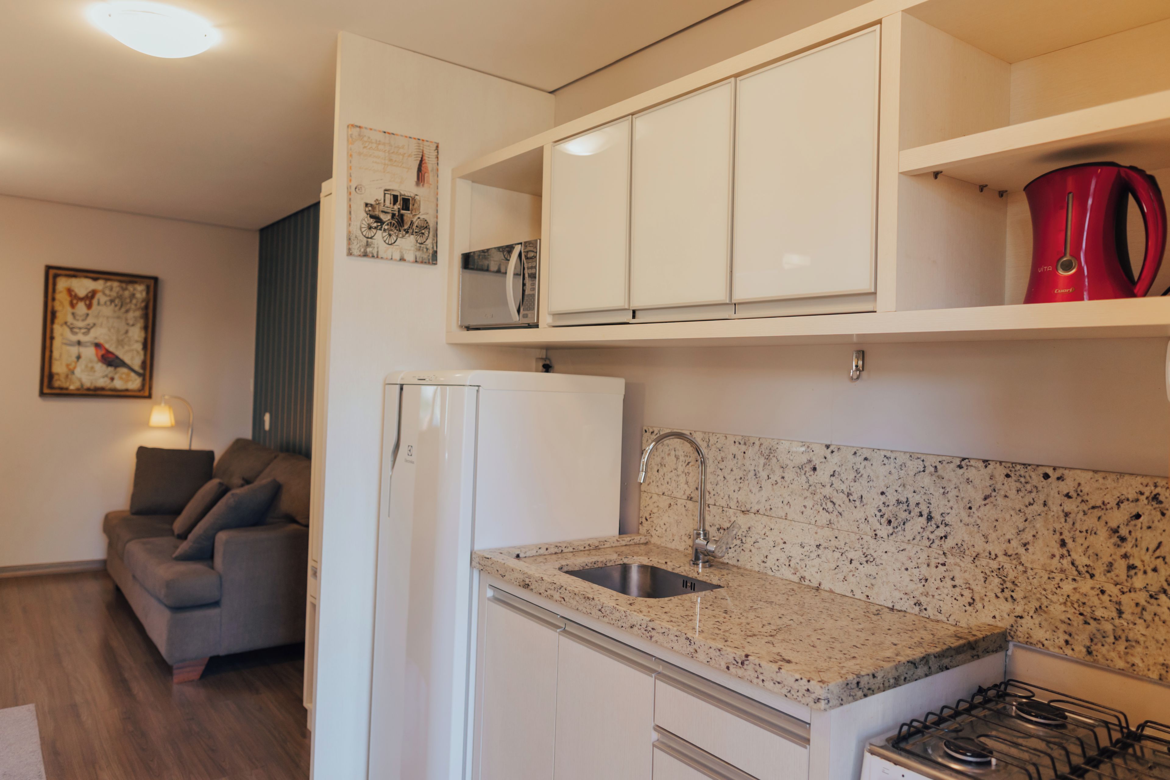 A compact kitchen with white cabinets, a stainless steel sink, and a red kettle on the countertop.