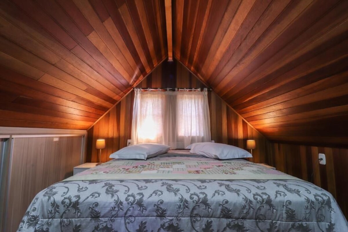 Quarto com teto inclinado, cama com colcha estampada, duas luminárias de cabeceira e janela com cortinas leves.
