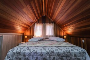 Un dormitorio con techo inclinado, una cama con colcha estampada, dos lámparas de noche y un ventanal con cortinas de gasa.